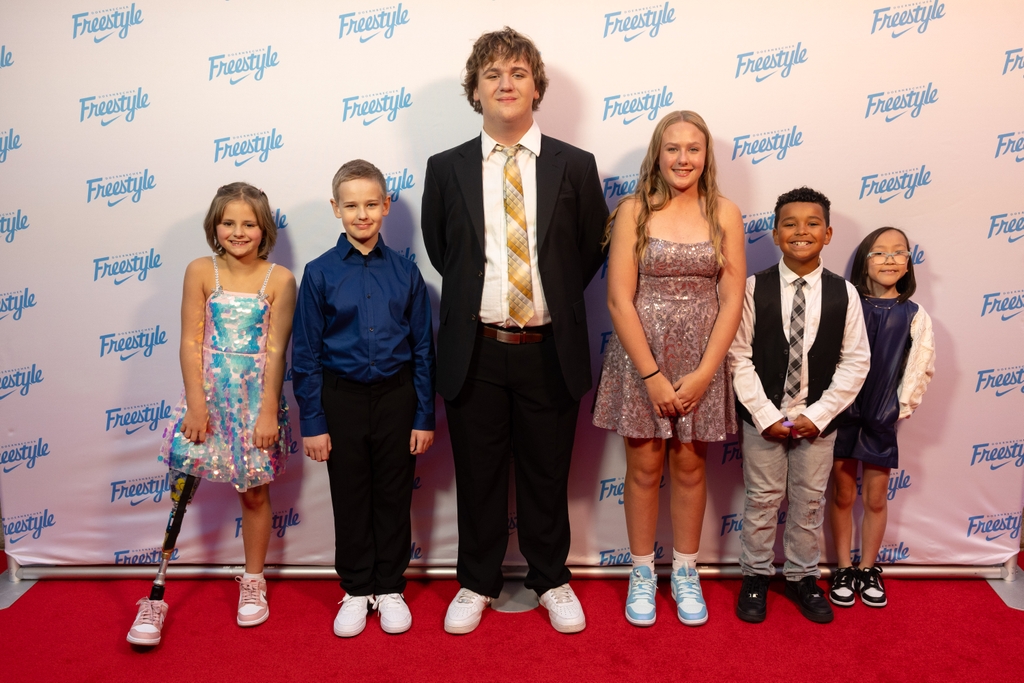Doernbecher Freestyle celebra 20 años. Seis jóvenes pacientes de OHSU Doernbecher revelaron sus zapatillas Nike diseñadas personalmente el viernes 18 de octubre de 2024. De izquierda a derecha: una niña con el pelo rubio corto recogido hacia atrás y un vestido de lentejuelas iridiscentes; un chico de cabello rubio corto y camisa de vestir azul oscuro y pantalón negro; un chico más alto con cabello corto y rubio y rizado, un traje negro con camisa de cuello blanco y corbata dorada; una chica de largo cabello rubio y un vestido de lentejuelas rosa claro; un niño de cabello corto oscuro y ondulado, pantalón gris, camisa de vestir blanca, chaleco negro y corbata; una chica con un peinado bob negro liso, anteojos y un vestido morado, todas con zapatillas de deporte. (Fundación OHSU/Fontaine Kaufman)