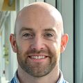 Jonathan Elliott, Ph.D., has no hair, a blue dress shirt, smiling in a hallway.