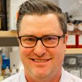 Benjamin Burwitz, Ph.D., has short brown hair, parted to the side, black-framed eye glasses and a white dress shirt, standing in a lab bay.