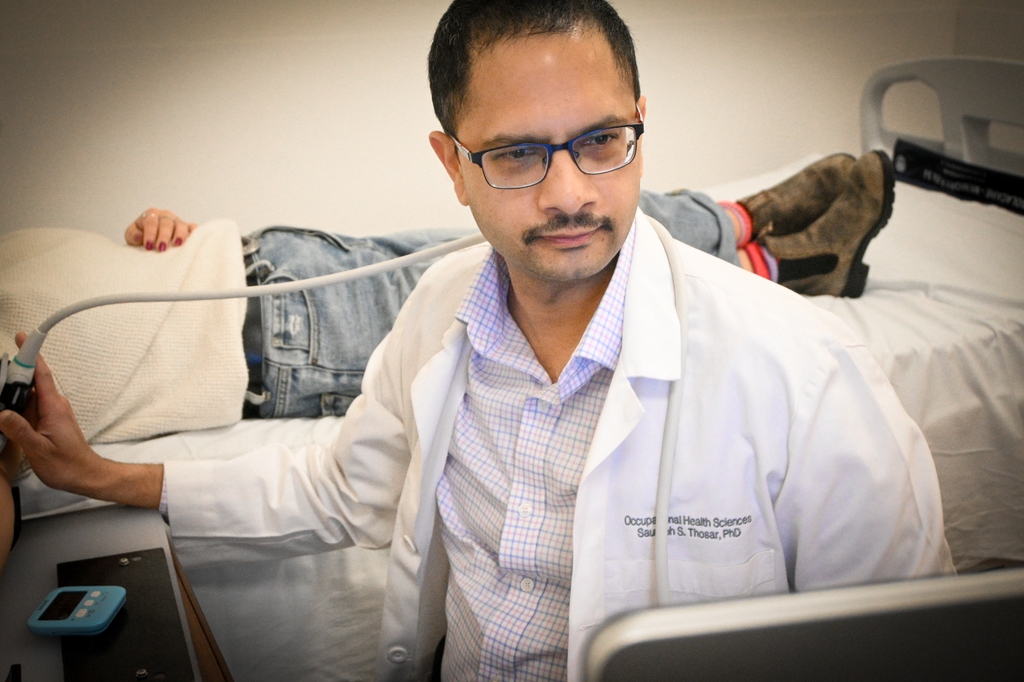 Saurabh Thosar, Ph.D., has short black hair, a mustache, eye glasses, wearing a white coat, and is smiling in a sleep study room. Saurabh Thosar, Ph.D., is leading new research at Oregon Health & Science University investigating the connection between sleep and cardiovascular health. (OHSU/Christine Torres Hicks)