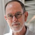 Michael Riscoe, Ph.D., tiene cabello corto gris oscuro, anteojos, es alto y viste una camisa abotonada de color azul claro, está de pie sobre un puente de conexión interior en OHSU.