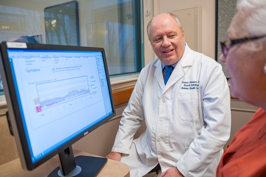 Andrw Ahmann has sparse white hair and wearing a white coat, talking with a patient while looking over diabetes data on a computer screen.