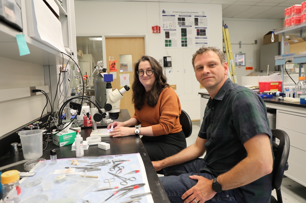 Jennifer Jahncke, on left, sits next to a microscope. She is wearing an orange top and eye glasses and has long wavy brown hair. On the right is Kevin Wright who has a button-up dark top on and has short wavy blond/brown hair.