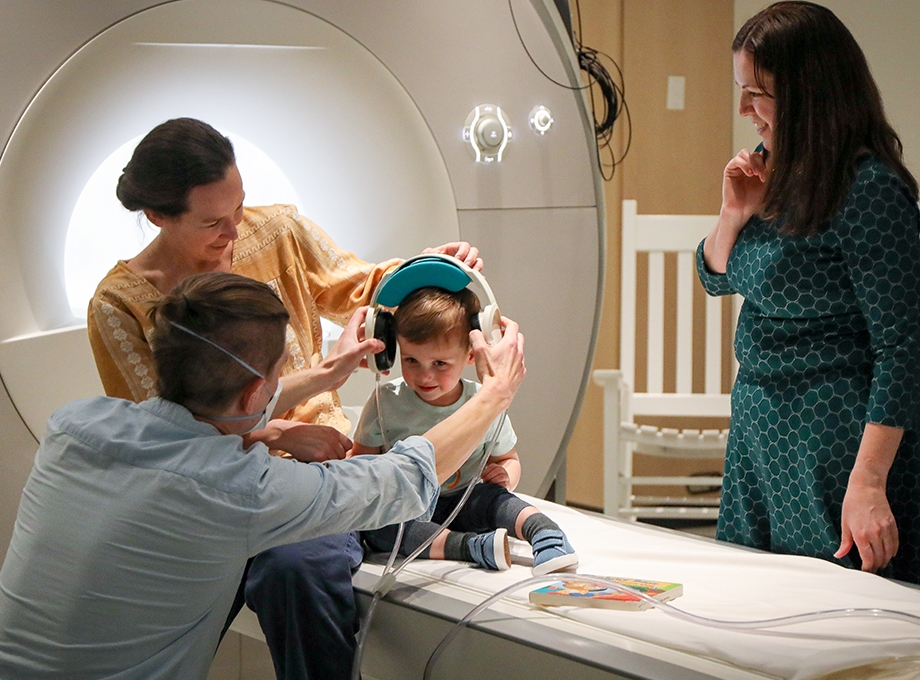 Estudio de audición pediátrico: La fotografía muestra a un niño pequeño sentado en una cama de hospital sonriendo mientras los investigadores le ponen auriculares en la cabeza.