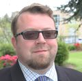 Alex Cotgreave has brown hair, parted to one side, dark sun glasses, and a suit, smiling in a garden area at OHSU.