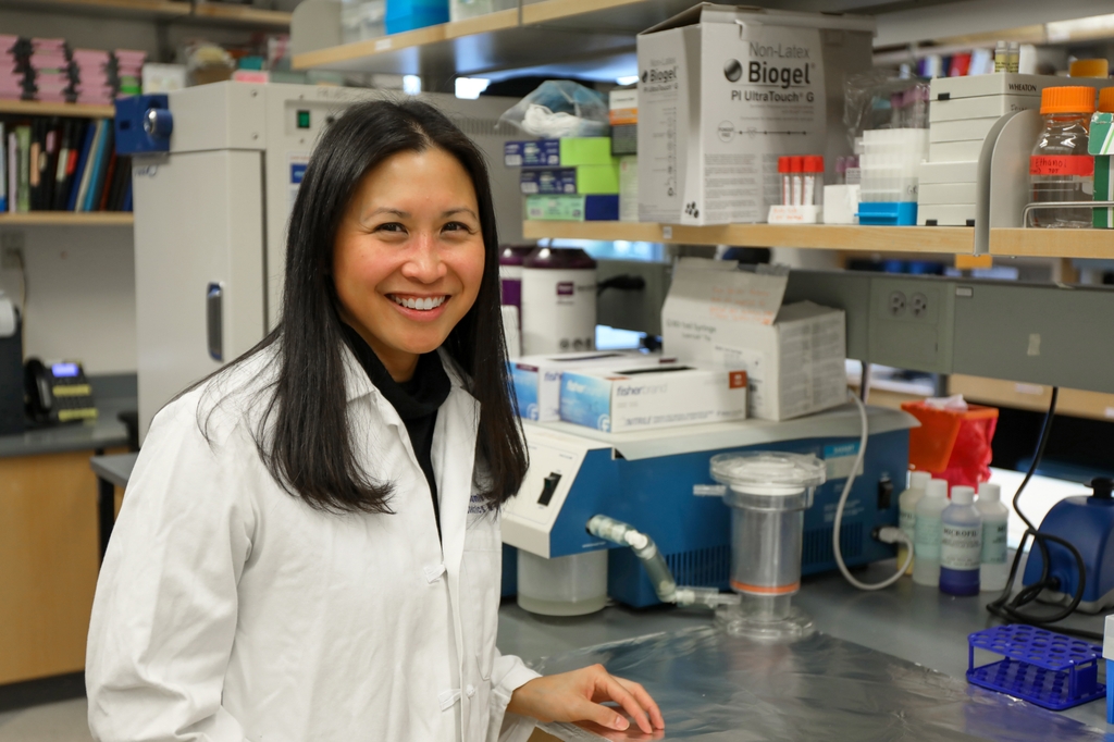 Jamie Lo, M.D., M.C.R. (OHSU) tiene cabello largo y oscuro y viste un suéter negro, parada en su laboratorio en OHSU.
