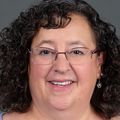 Jennifer Coury, M.S., has brown, tight curly hair, eye glasses and a light purple top, smiling.