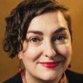 Lydia Bartholow, DNP, PMHNP, CARN-AP, FIAAN, has short dark curly hair, smiling against an orange background.
