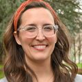 Elizabeth Wood, Ph.D., has long wavy brown hair, eye glasses, and an orange headband and gray top, smiling near OHSU fountain.