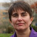 Nathalie Huguet, Ph.D., tiene cabello corto y oscuro, una camisa de vestir morada y sonríe cerca del Hospital OHSU (atrás).