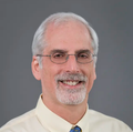 John Kaufman, M.D., M.S. (OHSU), in a dress shirt, has facial hair and gray hair, and glasses, smiling.