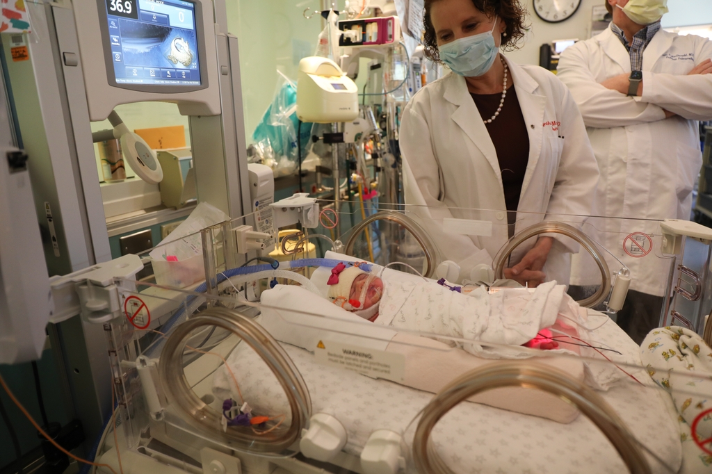 La imagen muestra a la Dra. Cindy McEvoy, con cabello corto y rizado, mascarilla y bata blanca, en la UCIN, supervisando una cuna con un bebé prematuro usando una CPAP. Un ensayo clínico realizado en la unidad de cuidados intensivos neonatales de OHSU Doernbecher ha identificado un nuevo protocolo de tratamiento para mejorar el crecimiento y la función pulmonar en bebés prematuros. La investigadora principal del estudio, la Dra. Cindy McEvoy, supervisa a un bebé prematuro usando terapia CPAP en la UCIN. (OHSU/Christine Torres Hicks)