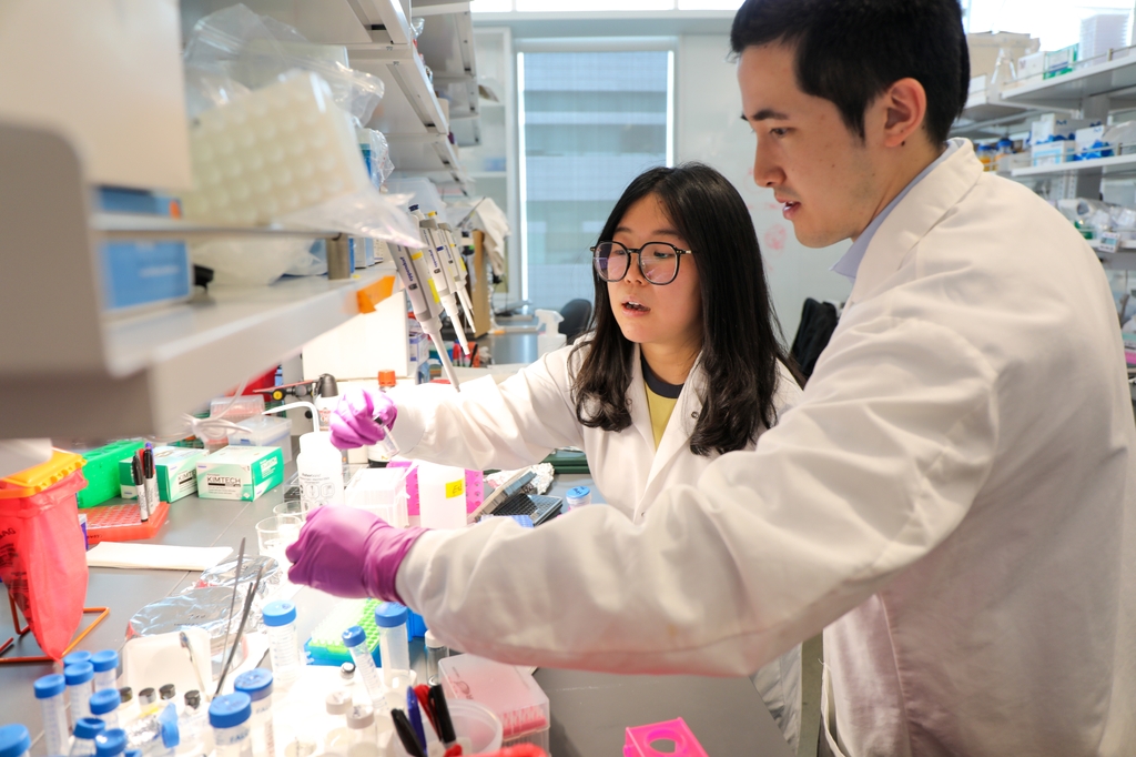 La imagen muestra a Li Xiang, de cabello largo y negro, gafas y bata blanca, mientras sostiene compuestos en un banco de investigación junto a Michael Henderson, de cabello oscuro y bata blanca. Li Xiang, doctora e investigadora postdoctoral en el Instituto Oncológico Knight de OHSU, y Michael Henderson, licenciado en Ingeniería Biomédica, forman parte de un equipo que desarrolló una nueva nanopartícula que aumenta la seguridad y eficacia del tratamiento del cáncer. (OHSU/Christine Torres Hicks)