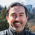 Alex Ortega Loayza, M.D., M.C.R. tiene el pelo oscuro partido, gafas y viste una camisa negra con un chaleco negro, sonriendo en la terraza del edificio OHSU Waterfront CHH1.