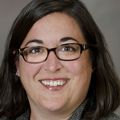 Erin Maynard has shoulder-length dark hair, eye glasses, gray suit and smiling against a gray background.