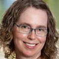 Shelley Tworoger, Ph.D. has curly long blonde hair, eye glasses, a red top, black jacket and smiling in a hallway.