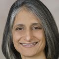Sima Desai has bobbed black and gray hair, a black top, smiling against a light tan background.
