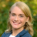 Michelle Hribar has wavy, medium blonde hair, smiling against a verdant background.