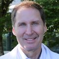 Timothy Smith, M.D., M.P.H. es alto, tiene cabello castaño claro/gris, ojos azules y viste una bata blanca mientras está de pie en un jardín en el Centro de Salud y Curación de OHSU.