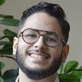 Vincenzo José Cornacchione Peña has short wavy black hair, eye glasses, facial hair and a gray/pink suit, smiling in a room with plants.