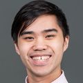 Bryan Le has short black hair and is wearing a light pink dress shirt, smiling in a photo studio that has a gray background.