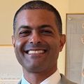 Shereef Elnahal, M.D., M.B.A., has shaved dark hair, is wearing a light blue suit with red tie and is smiling in a hallway.