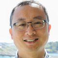 Zheng Xia, Ph.D., has very short dark hair, eye glasses, a blue/green shirt, and smiling with the Tillicum Bridge behind him.