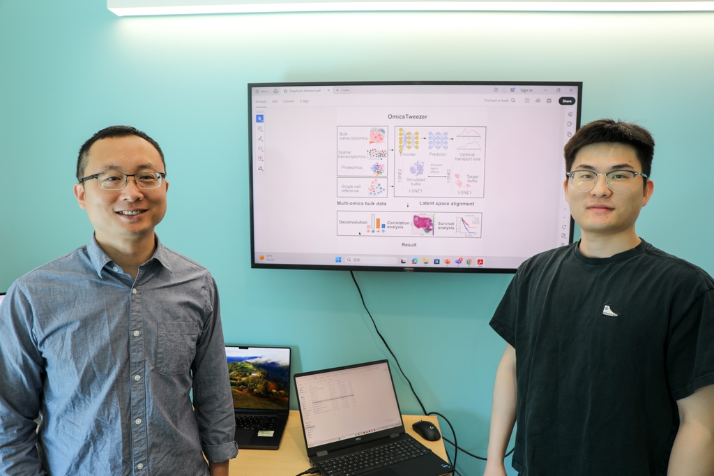 From left, Zheng Xia, Ph.D., has shaved short dark hair, eye glasses, and is wearing a blue/green shirt, standing next to a screen with a colorful data chart, and to the right, Xinxing Yang, Ph.D., who has short, dark hair, eye glasses, and a black t-shirt.