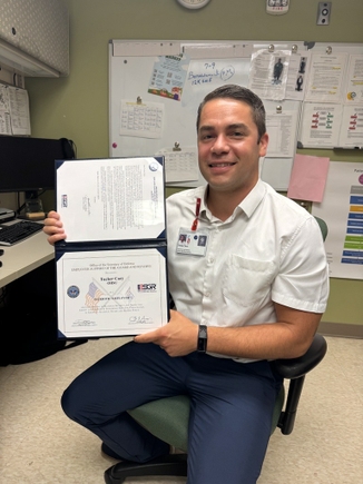 Tucker Cary has short dark gray/brown hair, a white shirt and blue pants, smiling while sitting in a chair in an office, holding his award.