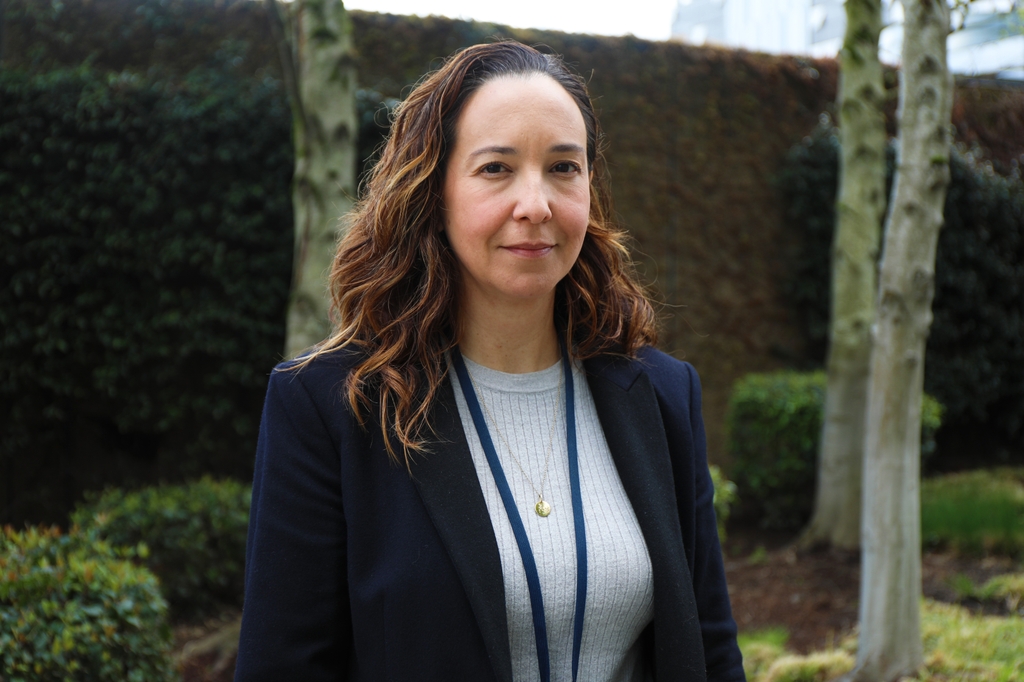 Image is of Ana, with long curly brown hair, a white top and black blazer, standing near greenery near OHSU CHH1. Ana Quiñones, Ph.D., is named co-director of the AGING Initiative, a $2.8 million grant from the NIH to advance a national research agenda focused on older adults with multiple chronic conditions. (OHSU/Christine Torres Hicks)