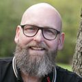 Marc Franke has no hair, facial hair, eye glasses, black shirt, smiling by a tree.