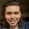 Adam Castillejo has long curly dark hair, a jacket and white shirt, smiling.