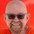 Paul Edmonds has an orange top, no hair, facial hair, dark sunglasses, smiling against an orange background