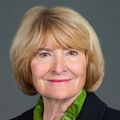 Susan King, M.S., RN, CEN, FAAN has a short bob blonde hair, black coat, green top and smiling against a gray background.