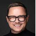 Justin Hurley Braswell has short brown hair, black glasses, black top, smiling.