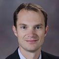 Brian Duty, M.D., M.B.A., has short brown hair, a suit on, and smiling against a grayish brown backdrop in a photo studio.