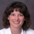 Kelly Anderson, FNP, has medium-length very curly brown hair, white clothes and smiling against a brown background in a photo studio.