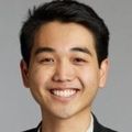 Nicholas Oshiro, M.H.A., has short, straight, side-parted black hair, a black coat and white shirt, smiling against a gray background in a photo studio.