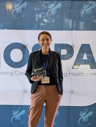 Kerith Hartmann, PA-C, has short cropped brown hair and a black jacket with brown pants, smiling while holding an award.