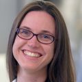 Julia has long straight brown hair, eye glasses and wearing a white coat, smiling in her lab.