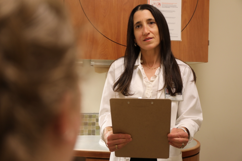 Cantor holds a clipboard, interviewing a patient in a patient room.