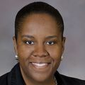 Monique has dark hair pulled back, a black suit, smiling in a photo studio.