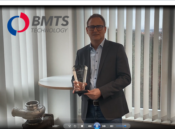 Smiling man in suit with glasses holding award next to exhaust turbocharger, in front of vertical blinds, logo superimposed