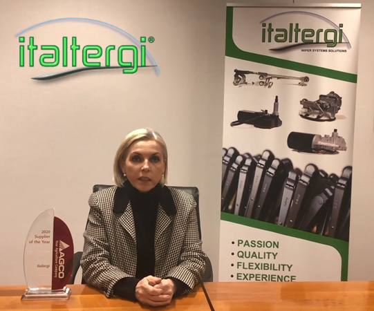Woman seated at desk with award placed on desk, sign next to her, logo superimposed
