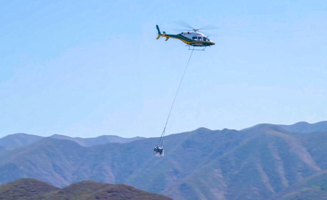 A reel of covered conductor is airlifted from the staging area to one of the remote pole locations where the insulated power lines will be strung using a helicopter. 