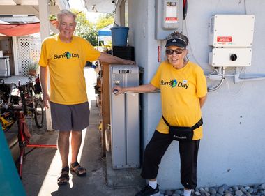 Two people in yellow "Sun Day" shirts stand outside, smiling near electrical equipment.