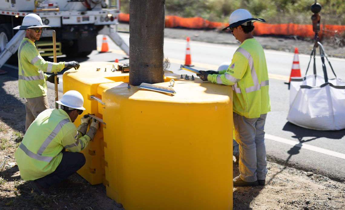 Workers in safety gear install a large yellow barrier on a roadside next to orange traffic cones and equipment.
