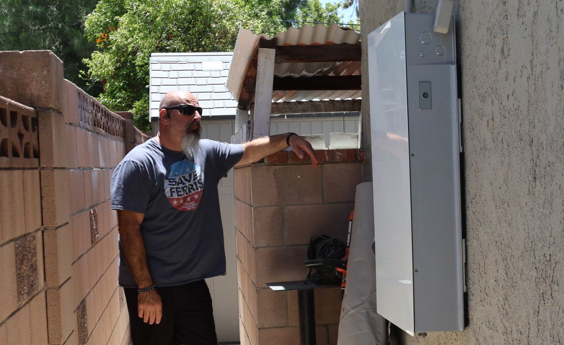 A man with a beard and sunglasses leans against a wall next to an outdoor, wall-mounted home battery.