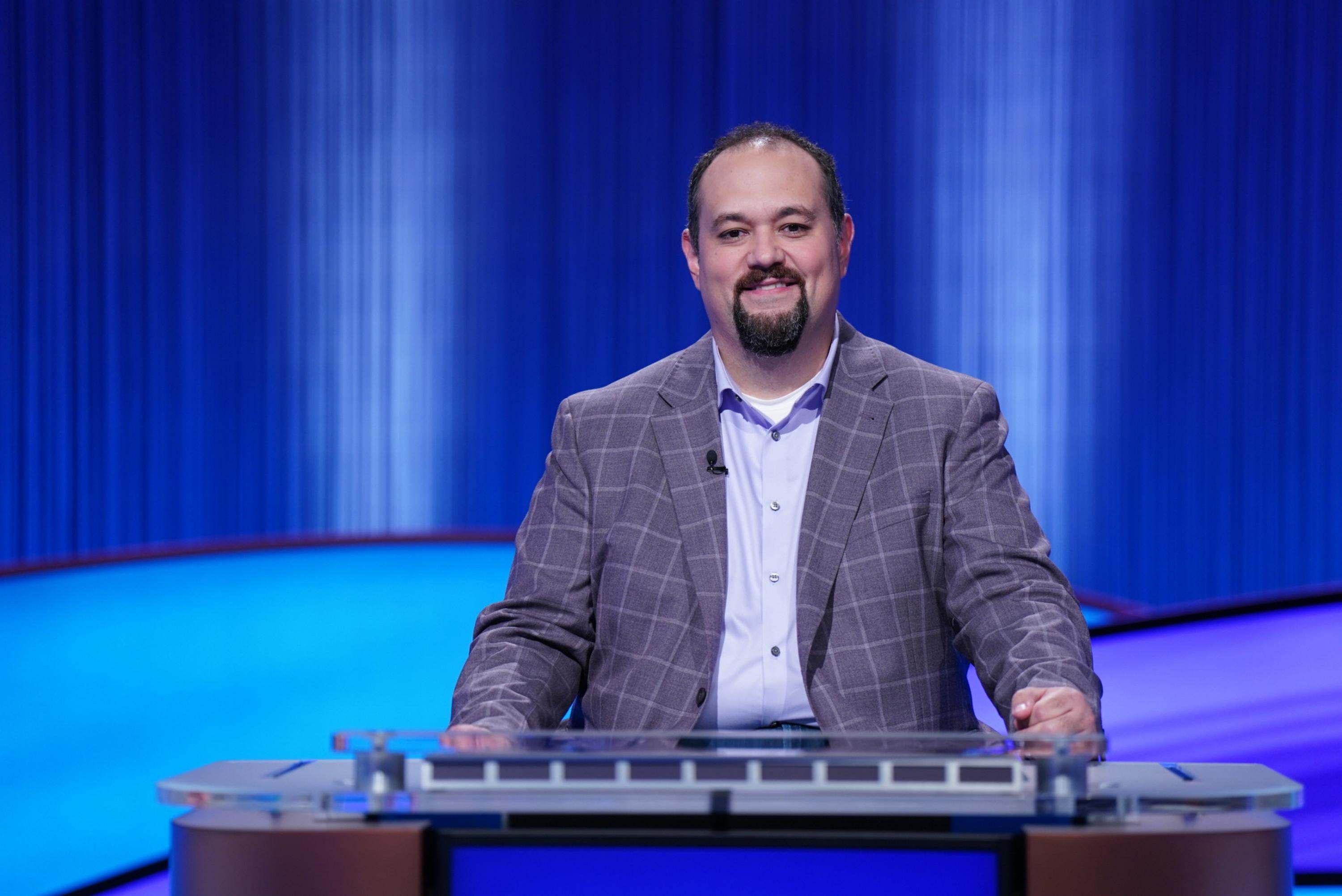 Man in a plaid suit stands behind a podium against a blue curtain background.