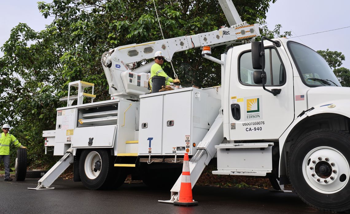 Utility workers operate a bucket truck on a road lined with trees, accompanied by traffic cones for safety.